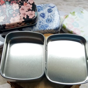 K&ouml;nnte beinhalten: Eine offene, silberfarbene Scharnierdose mit rechteckiger Form. Im Hintergrund sind mehrere andere dekorative Dosen mit Blumen- und Polka-Dot-Mustern zu sehen. Die Dosen sind klein und scheinen aus Metall zu sein.