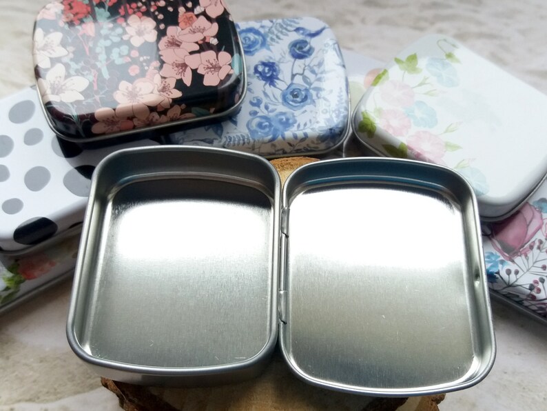 K&ouml;nnte beinhalten: Eine offene, silberfarbene Scharnierdose mit rechteckiger Form. Im Hintergrund sind mehrere andere dekorative Dosen mit Blumen- und Polka-Dot-Mustern zu sehen. Die Dosen sind klein und scheinen aus Metall zu sein.