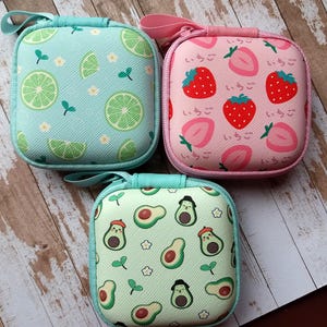 Puede incluir: Tres peque&ntilde;os estuches cuadrados con cremallera y estampados de frutas. Uno es azul claro con rodajas de lima, otro es rosa con fresas y texto japon&eacute;s, y el tercero es verde claro con personajes de aguacate.