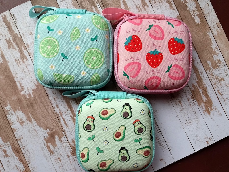 Puede incluir: Tres peque&ntilde;os estuches cuadrados con cremallera y estampados de frutas. Uno es azul claro con rodajas de lima, otro es rosa con fresas y texto japon&eacute;s, y el tercero es verde claro con personajes de aguacate.