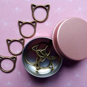 May include: A set of gold cat-shaped stitch markers in a pink metal tin. The tin is open and the markers are spilling out.