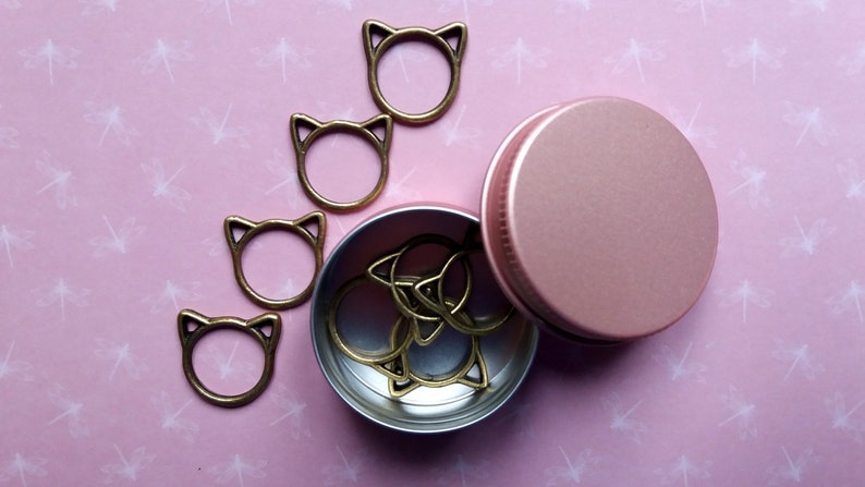 May include: A set of gold cat-shaped stitch markers in a pink metal tin. The tin is open and the markers are spilling out.