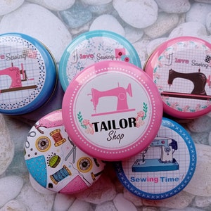 Op de afbeelding: Een verzameling ronde, decoratieve blikken met naaipatronen. De blikken tonen afbeeldingen van naaimachines, garenklosjes en de tekst "I Love Sewing" en "Tailor Shop". De blikken zijn in verschillende kleuren, waaronder blauw, roze en wit.