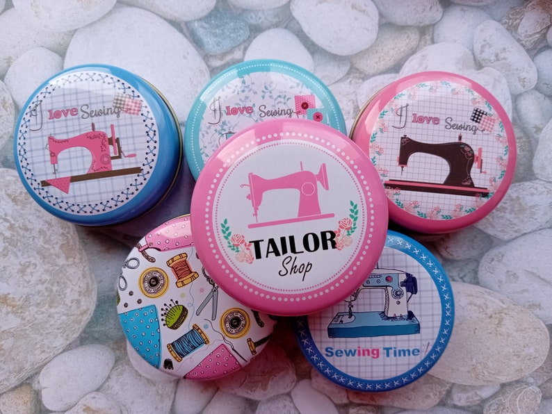 Op de afbeelding: Een verzameling ronde, decoratieve blikken met naaipatronen. De blikken tonen afbeeldingen van naaimachines, garenklosjes en de tekst "I Love Sewing" en "Tailor Shop". De blikken zijn in verschillende kleuren, waaronder blauw, roze en wit.