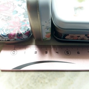 K&ouml;nnte beinhalten: Nahaufnahme von drei rechteckigen Metallboxen mit floralen Designs. Die Dosen sind wei&szlig; mit rosa, blauen und lila Blumen. Ein rosa Lineal mit schwarzen Markierungen befindet sich im Vordergrund und zeigt die Ma&szlig;e in Zentimetern an.