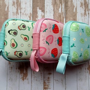 Puede incluir: Tres peque&ntilde;as bolsas cuadradas con cremallera y estampados de frutas. Una es verde claro con dise&ntilde;os de aguacate, otra es rosa con fresas y la tercera es azul claro con rodajas de lima. Cada una tiene una cremallera y una correa de mu&ntilde;eca del mismo color.