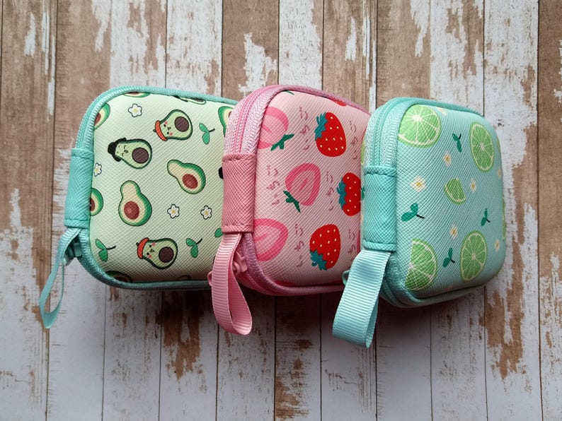 Puede incluir: Tres peque&ntilde;as bolsas cuadradas con cremallera y estampados de frutas. Una es verde claro con dise&ntilde;os de aguacate, otra es rosa con fresas y la tercera es azul claro con rodajas de lima. Cada una tiene una cremallera y una correa de mu&ntilde;eca del mismo color.
