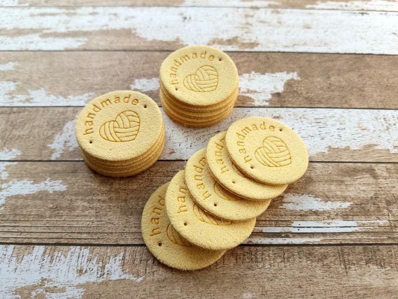 May include: A stack of 10 yellow round leather tags with the words "handmade" and a heart with yarn inside.