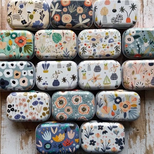 Pu&ograve; includere: Una collezione di scatole metalliche rettangolari con disegni floreali e botanici. Le scatole presentano varie palette di colori, tra cui blu, verdi e arancioni, con motivi di fiori, foglie e forme astratte. Le scatole sono disposte su una superficie di legno.