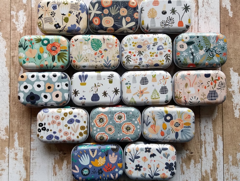 Pu&ograve; includere: Una collezione di scatole metalliche rettangolari con disegni floreali e botanici. Le scatole presentano varie palette di colori, tra cui blu, verdi e arancioni, con motivi di fiori, foglie e forme astratte. Le scatole sono disposte su una superficie di legno.