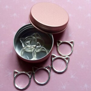 May include: A pink metal container with a lid, filled with silver cat-shaped stitch markers.  There are also several cat-shaped stitch markers outside of the container on a pink background.