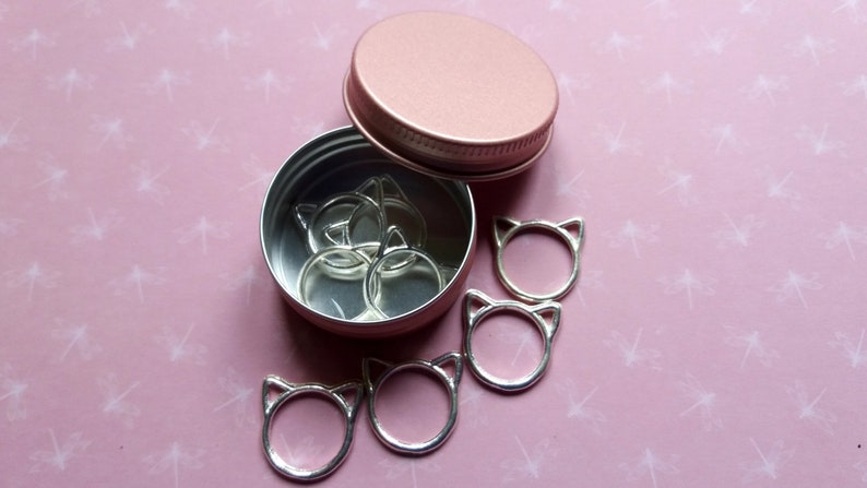 May include: A pink metal container with a lid, filled with silver cat-shaped stitch markers.  There are also several cat-shaped stitch markers outside of the container on a pink background.