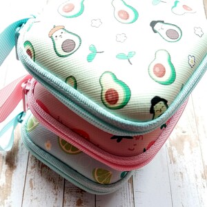 Puede incluir: Tres estuches peque&ntilde;os con cremallera apilados. La funda superior es blanca con dibujos de aguacates verdes. La funda del medio es rosa y la inferior azul claro con un dise&ntilde;o de lima. Cada estuche tiene una cremallera y un asa del mismo color.