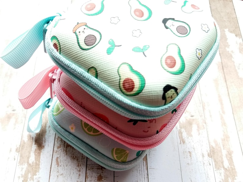 Puede incluir: Tres estuches peque&ntilde;os con cremallera apilados. La funda superior es blanca con dibujos de aguacates verdes. La funda del medio es rosa y la inferior azul claro con un dise&ntilde;o de lima. Cada estuche tiene una cremallera y un asa del mismo color.