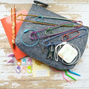 Könnte beinhalten: Ein graues Filz-Strickset mit verschiedenen Werkzeugen, darunter große Maschenhalter, eine kleine Schere und ein Maßband. Ein floraler Stift und ein orangefarbener Notizblock sind ebenfalls vorhanden, zusammen mit farbenfrohen Maschenmarkierern und einer kleinen Karte mit Text.