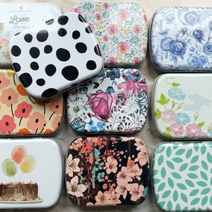 K&ouml;nnte beinhalten: Eine Sammlung rechteckiger Metalldosen mit verschiedenen Designs. Die Dosen zeigen florale Muster, Polka Dots und Illustrationen von Luftballons, einem Kuchen und einem Vogel. Die Farbpalette umfasst Pastellt&ouml;ne, Schwarz und Wei&szlig;.