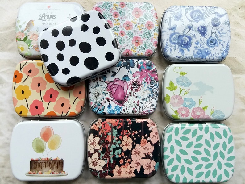 K&ouml;nnte beinhalten: Eine Sammlung rechteckiger Metalldosen mit verschiedenen Designs. Die Dosen zeigen florale Muster, Polka Dots und Illustrationen von Luftballons, einem Kuchen und einem Vogel. Die Farbpalette umfasst Pastellt&ouml;ne, Schwarz und Wei&szlig;.