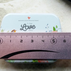 K&ouml;nnte beinhalten: Eine rechteckige, hellblaue Metallbox mit floralem Design und dem Text "A true Love story". Die Box ist etwa 5 cm breit. Der Deckel zeigt einen Vogel und ein Herz.