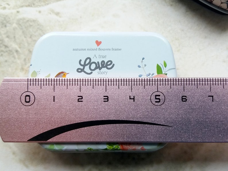 K&ouml;nnte beinhalten: Eine rechteckige, hellblaue Metallbox mit floralem Design und dem Text "A true Love story". Die Box ist etwa 5 cm breit. Der Deckel zeigt einen Vogel und ein Herz.