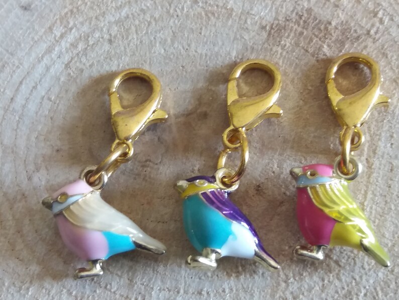 May include: Three gold-tone bird charms with colorful enamel paint. Each charm features a different color combination of pink, blue, yellow, white, and purple. The charms have a lobster claw clasp.