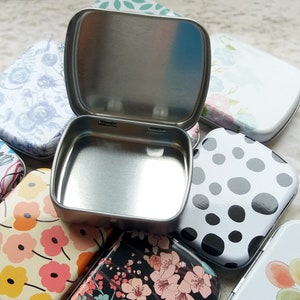 K&ouml;nnte beinhalten: Eine offene, silberfarbene Metallbox liegt inmitten mehrerer geschlossener Dosen. Die geschlossenen Dosen weisen verschiedene Blumen- und Polka-Dot-Muster in Farben wie Rosa, Wei&szlig; und Schwarz auf. Die offene Dose ist quadratisch mit abgerundeten Ecken.