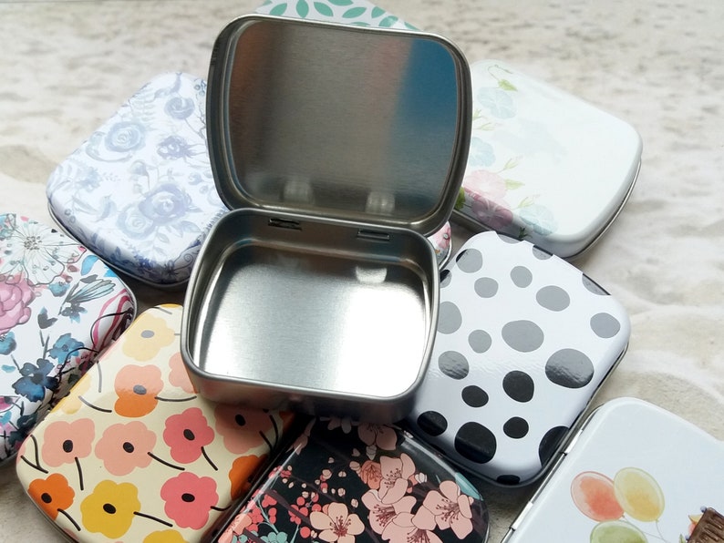 K&ouml;nnte beinhalten: Eine offene, silberfarbene Metallbox liegt inmitten mehrerer geschlossener Dosen. Die geschlossenen Dosen weisen verschiedene Blumen- und Polka-Dot-Muster in Farben wie Rosa, Wei&szlig; und Schwarz auf. Die offene Dose ist quadratisch mit abgerundeten Ecken.