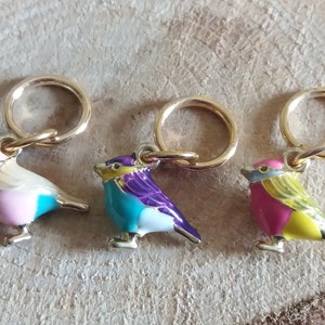 May include: Three colorful bird-shaped stitch markers with gold rings. The birds are pink and blue, turquoise and purple, and pink and yellow.