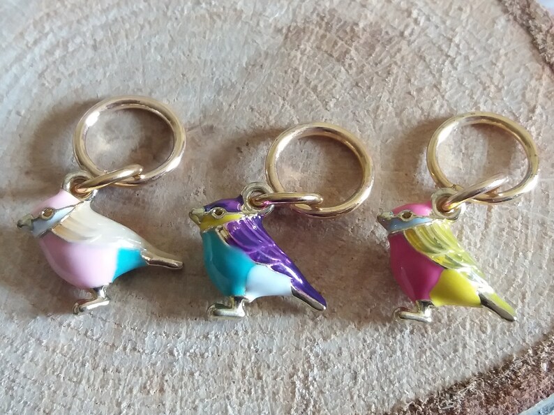 May include: Three colorful bird-shaped stitch markers with gold rings. The birds are pink and blue, turquoise and purple, and pink and yellow.
