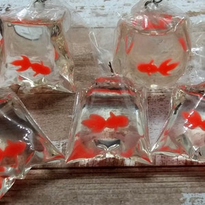 K&ouml;nnte beinhalten: F&uuml;nf transparente Plastikt&uuml;ten mit jeweils einem leuchtend orangefarbenen Goldfisch. Die T&uuml;ten sind oben mit einem silberfarbenen Draht verschlossen. Die Goldfische sind in den T&uuml;ten sichtbar, die auf einer Holzoberfl&auml;che angeordnet sind.
