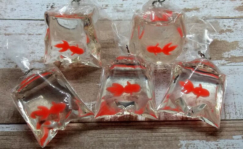 K&ouml;nnte beinhalten: F&uuml;nf transparente Plastikt&uuml;ten mit jeweils einem leuchtend orangefarbenen Goldfisch. Die T&uuml;ten sind oben mit einem silberfarbenen Draht verschlossen. Die Goldfische sind in den T&uuml;ten sichtbar, die auf einer Holzoberfl&auml;che angeordnet sind.