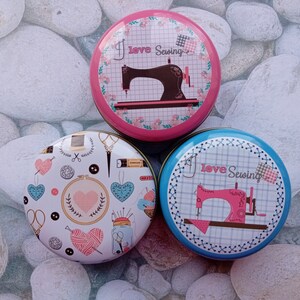 Op de afbeelding: Drie ronde naaidozen met kleurrijke ontwerpen. E&eacute;n doos heeft een roze achtergrond met een naaimachine-afbeelding en de tekst "I love Sewing". Een andere doos heeft een blauwe achtergrond met dezelfde tekst en een roze naaimachine. De derde doos heeft een witte achtergrond met naaigerelateerde illustraties.