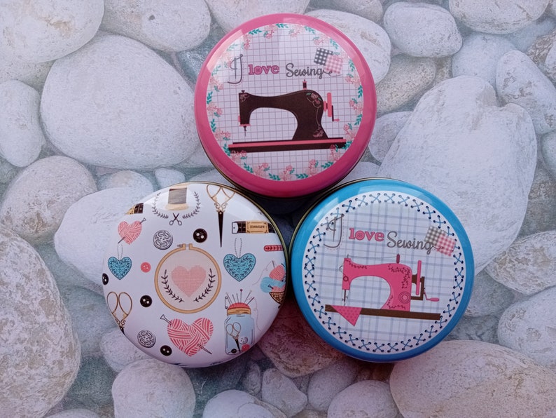 Op de afbeelding: Drie ronde naaidozen met kleurrijke ontwerpen. E&eacute;n doos heeft een roze achtergrond met een naaimachine-afbeelding en de tekst "I love Sewing". Een andere doos heeft een blauwe achtergrond met dezelfde tekst en een roze naaimachine. De derde doos heeft een witte achtergrond met naaigerelateerde illustraties.