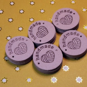 May include: Six purple round leather tags with the text "handmade" and a heart with a yarn design. The tags are on a gold background with white stars.