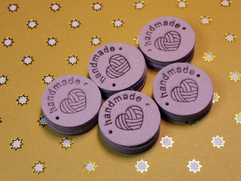 May include: Six purple round leather tags with the text "handmade" and a heart with a yarn design. The tags are on a gold background with white stars.