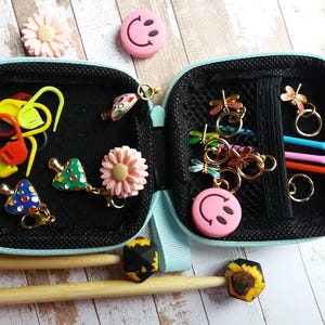 Puede incluir: Estuche negro abierto con cremallera con varios accesorios de tejido. Incluye marcadores de punto, dijes de lib&eacute;lula, dijes de carita sonriente rosa y ganchillos. Tambi&eacute;n son visibles dijes decorativos y agujas de tejer de madera.