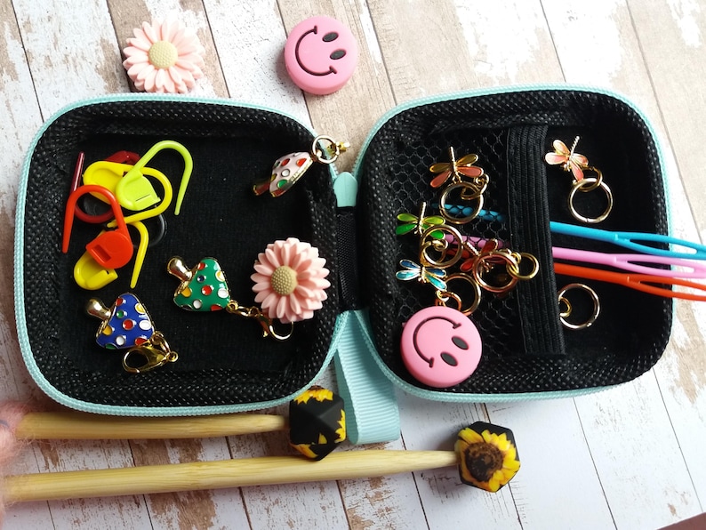 Puede incluir: Estuche negro abierto con cremallera con varios accesorios de tejido. Incluye marcadores de punto, dijes de lib&eacute;lula, dijes de carita sonriente rosa y ganchillos. Tambi&eacute;n son visibles dijes decorativos y agujas de tejer de madera.