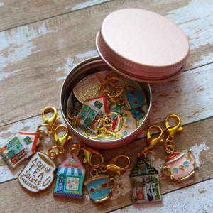 May include: A pink metal container with a lid filled with colorful enamel charms. The charms are shaped like houses, teacups, and teapots. The charms have gold metal rings and clasps. The text on one charm reads "A Cup of Tea Solves Everything".