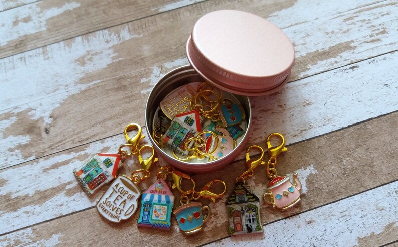 May include: A pink metal container with a lid filled with colorful enamel charms. The charms are shaped like houses, teacups, and teapots. The charms have gold metal rings and clasps. The text on one charm reads "A Cup of Tea Solves Everything".