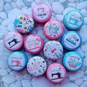 Op de afbeelding: Een verzameling ronde, decoratieve blikken met naaigerelateerde ontwerpen. De blikken tonen afbeeldingen van naaimachines, garenklosjes, scharen en de tekst "TAILOR Shop", "I love Sewing" en "Sewing Time". De blikken zijn in verschillende kleuren, waaronder roze, blauw en wit.