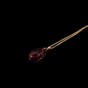 Pode incluir: Um colar de corrente dourada com um pingente grande em forma de gota. O pingente é de cor vermelha escura com manchas douradas. O colar está sobre um fundo preto.