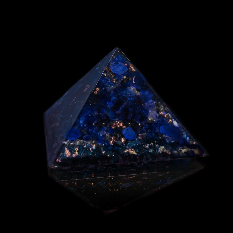 May include: A dark blue pyramid-shaped object with embedded blue and gold-colored elements. The pyramid is set against a black background, with a reflective surface below. The object appears to be made of resin or a similar material.