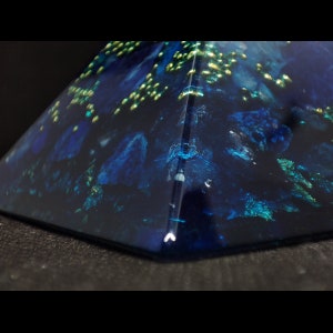 May include: A close-up of a pyramid-shaped object with a dark blue, almost black, base. The pyramid has a glossy finish with embedded blue and green accents, and small gold flecks. The object is set against a dark background.