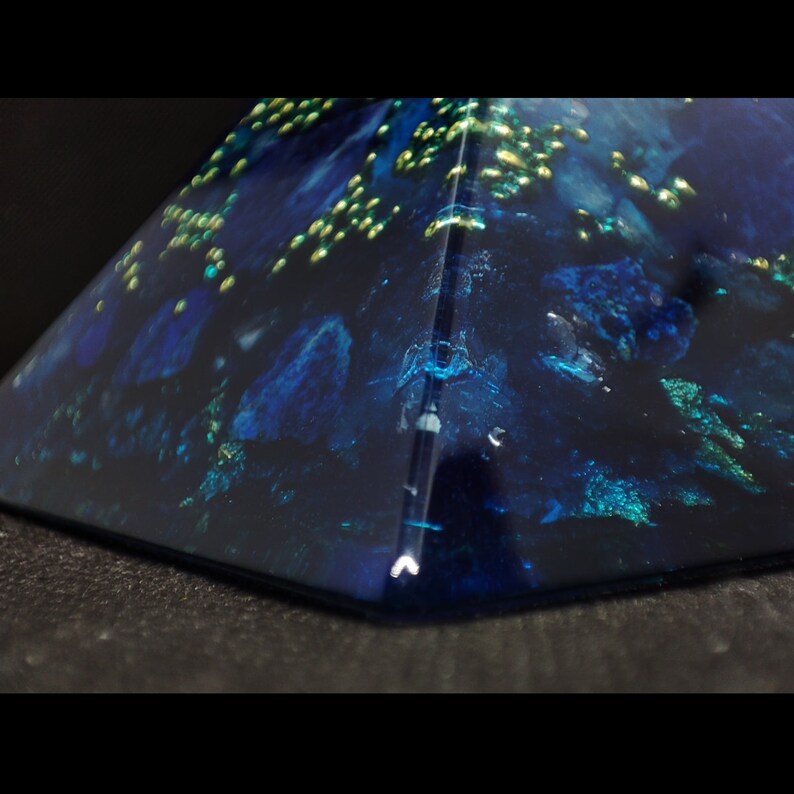 May include: A close-up of a pyramid-shaped object with a dark blue, almost black, base. The pyramid has a glossy finish with embedded blue and green accents, and small gold flecks. The object is set against a dark background.