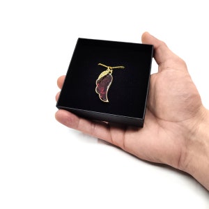 Puede incluir: Un collar dorado con un colgante rojo y dorado en forma de ala, presentado en una caja negra. El colgante tiene un contorno dorado y un interior rojo texturizado. El collar está en una cadena dorada.