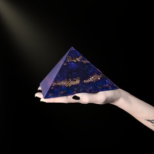May include: A dark blue pyramid with gold accents is held in a hand against a black background. The pyramid has a textured surface with embedded gold spheres and a subtle sparkle. The pyramid shape is a geometric design.