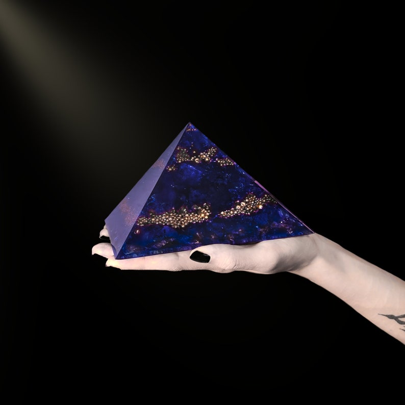 May include: A dark blue pyramid with gold accents is held in a hand against a black background. The pyramid has a textured surface with embedded gold spheres and a subtle sparkle. The pyramid shape is a geometric design.