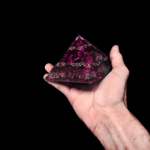 Könnte beinhalten: Handgefertigte, pyramidenförmige Dekoration in dunklem Lila, eingebettet mit silberfarbenen Perlen und anderen dekorativen Elementen. Die Pyramide wird in einer Hand vor schwarzem Hintergrund gehalten, wodurch ihre geometrische Form und die feinen Details hervorgehoben werden.