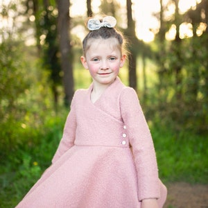 May include: A young girl wearing a pink dress with a white bow in her hair. She is standing in a forest setting with green trees and grass.