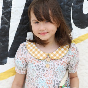 May include: A young girl wearing a floral dress with a yellow and white gingham collar and holding a book. She is standing in front of a white wall with black and yellow graffiti.