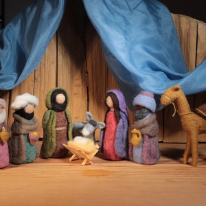 May include: A felt nativity scene with figures of the Holy Family, wise men, a donkey, and a camel. The figures are in various colours, including purple, green, and brown, set against a wooden backdrop with blue fabric.